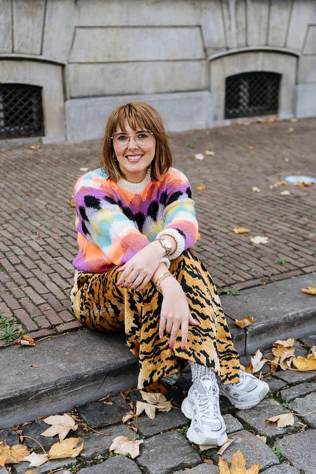 Vrouw in kleurrijke trui en broek met dierenprint, lachend buiten zittend op de stoeprand.