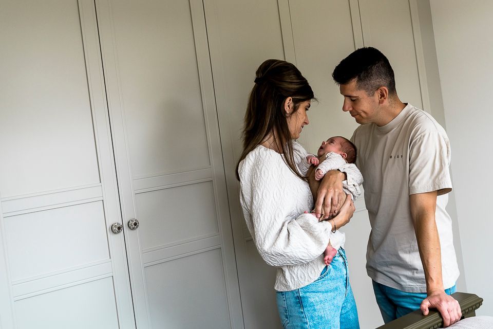 Een stel kijkt vol bewondering naar hun pasgeboren baby, die ze in hun armen houden, voor een witte kast.
