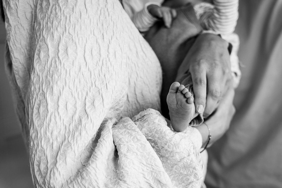 Persoon die een pasgeboren baby vasthoudt; details zijn onder andere kleine voetjes, een stof met textuur en grijstinten.