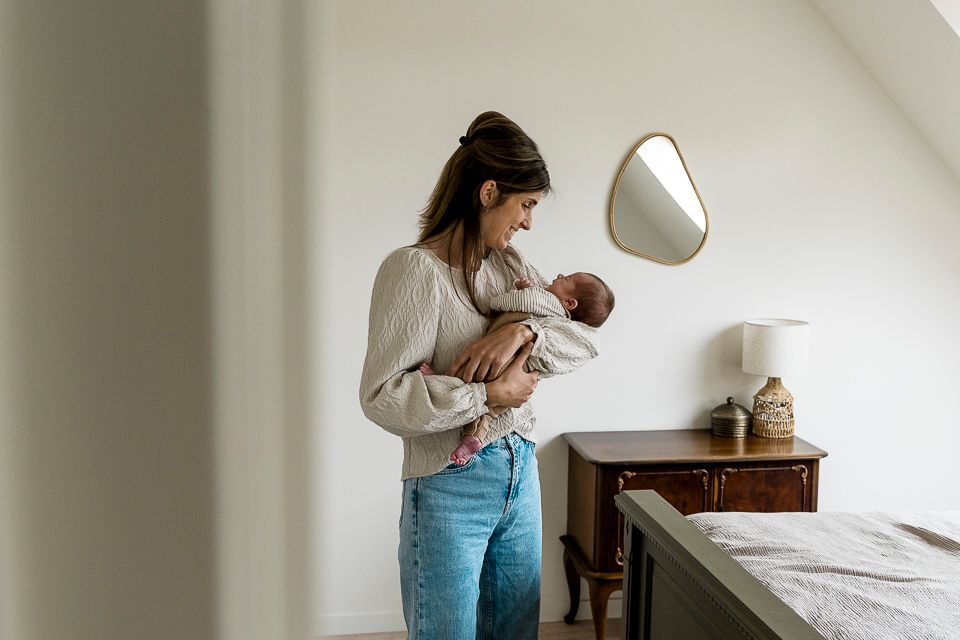 Vrouw houdt een pasgeboren baby vast in een slaapkamer en kijkt naar beneden. Neutrale kleuren, met een spiegel en nachtkastje zichtbaar.