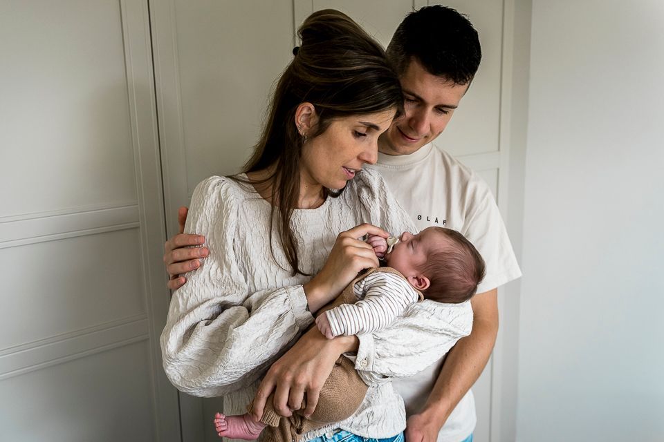 Ouders houden een pasgeboren baby vast en kijken ernaar binnenshuis, vlakbij een witte kast.