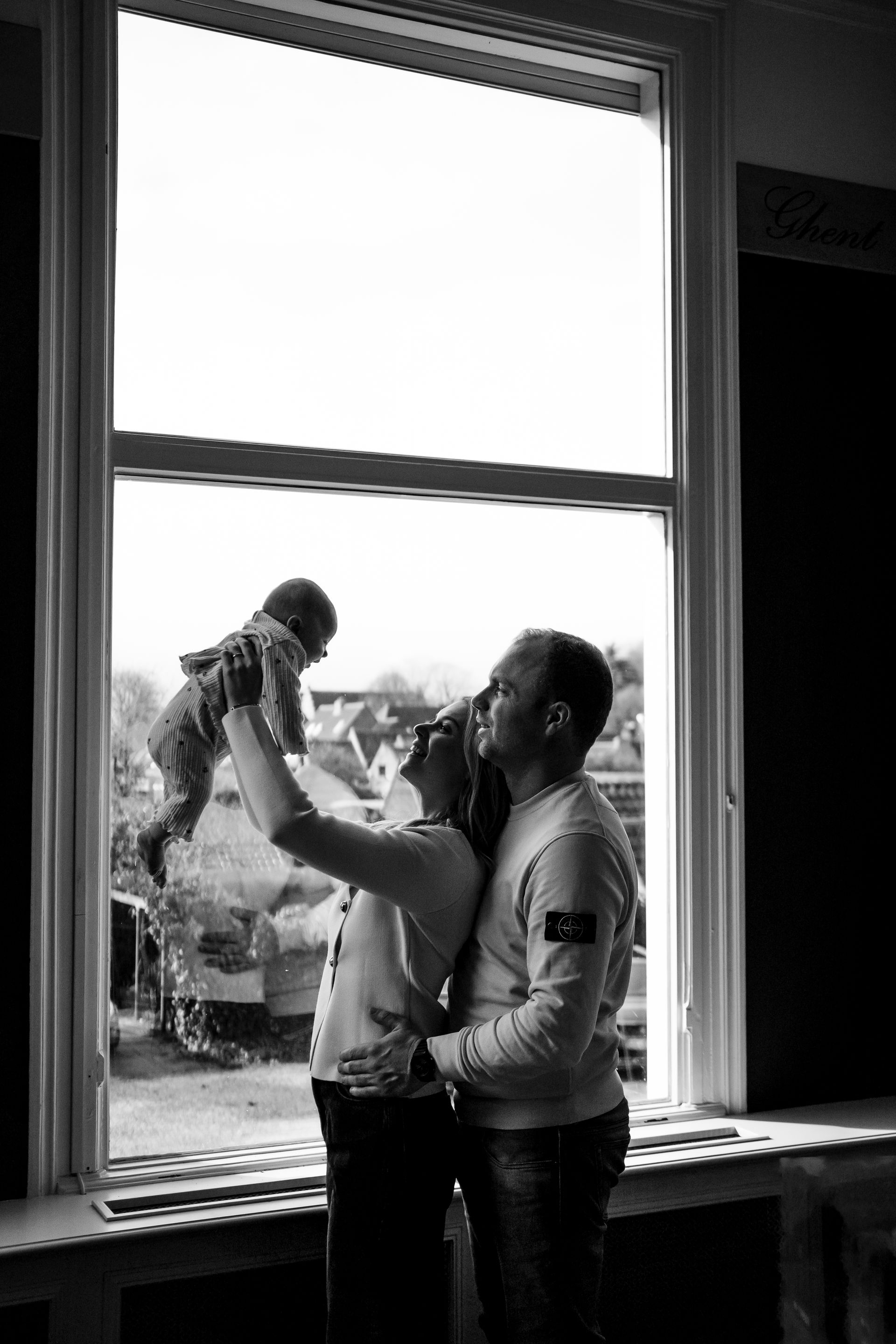 Een gezin zit binnen bij een raam en houdt een baby vast. Het silhouet is te zien tegen het buitenlicht.