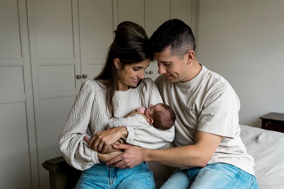 Ouders kijken naar hun pasgeboren baby, binnenshuis. Moeder houdt de baby vast, vader kijkt toe. Warme tinten, neutrale omgeving.
