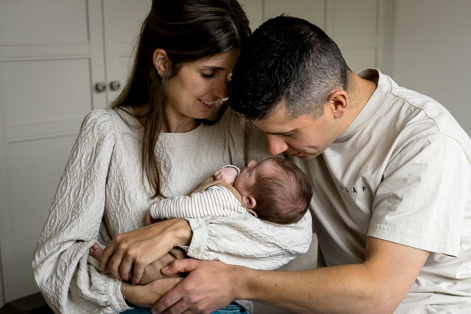 Ouders houden hun pasgeboren baby vast, binnenshuis. Moeder kijkt naar beneden en glimlacht; vader kust het hoofdje van de baby.