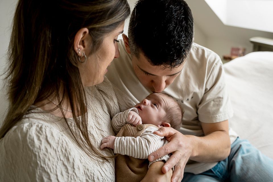 Ouders knuffelen een pasgeboren baby, moeder kijkt toe, vader kust het hoofdje van de baby, binnen, zacht licht.