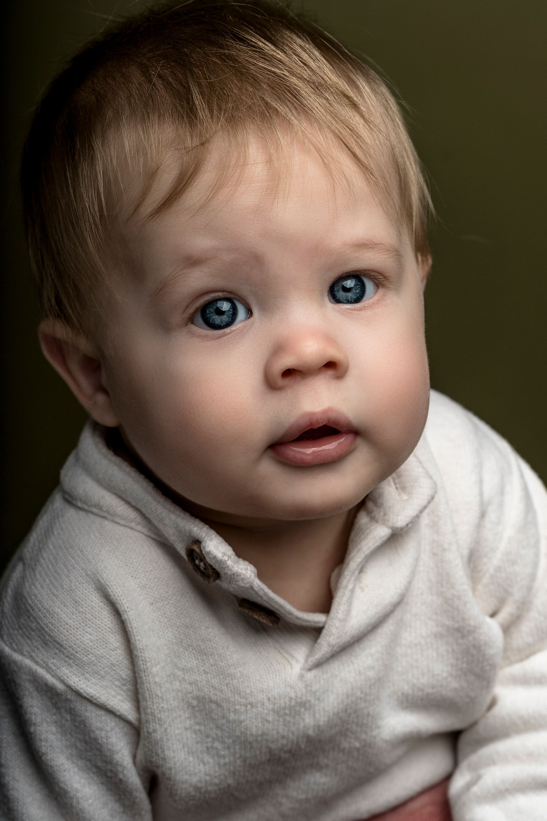 Een baby met blond haar en blauwe ogen, gekleed in een witte trui, kijkt aandachtig naar de kijker.