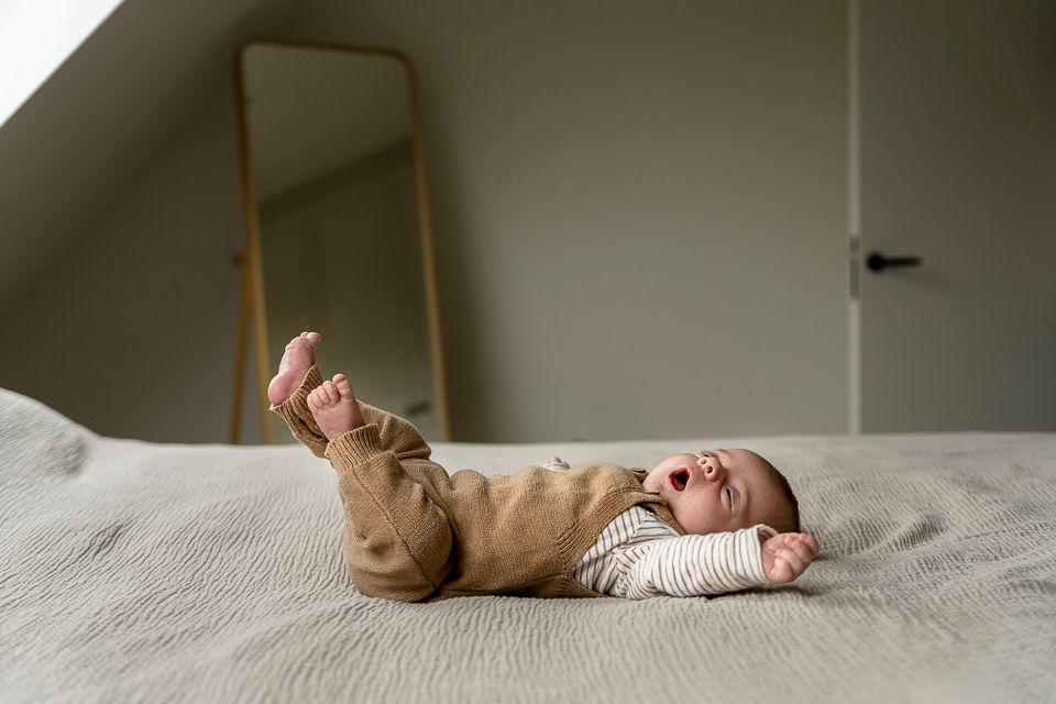 Een baby gaapt op een bed, gekleed in een bruine tuinbroek en een gestreept shirt, met de armen en voeten in de lucht.