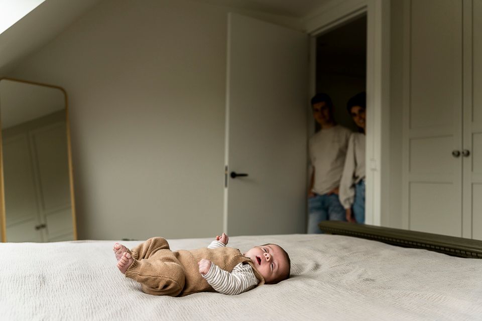 Een baby ligt op een bed en kijkt omhoog. Op de achtergrond staan twee mensen in een deuropening en kijken toe.
