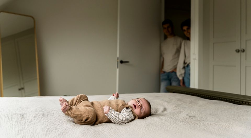 Baby ligt in bed, ouders kijken toe vanuit de deuropening. Lichte kamer met een spiegel en neutrale kleuren.