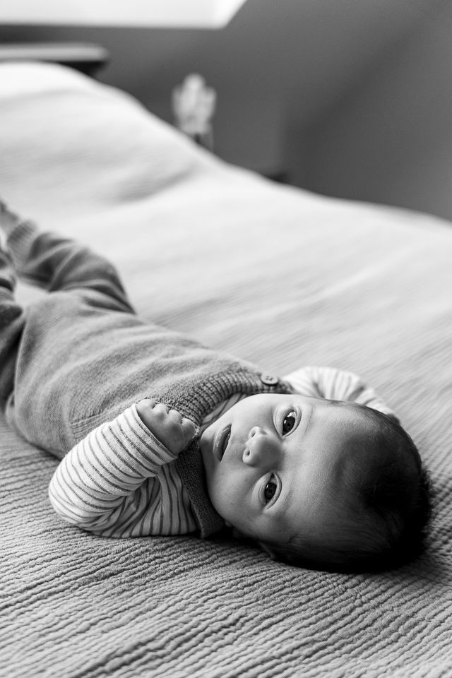 Baby ligt op bed en kijkt naar de camera. Draagt een gestreept shirt en een tuinbroek. Zwart-witfoto.