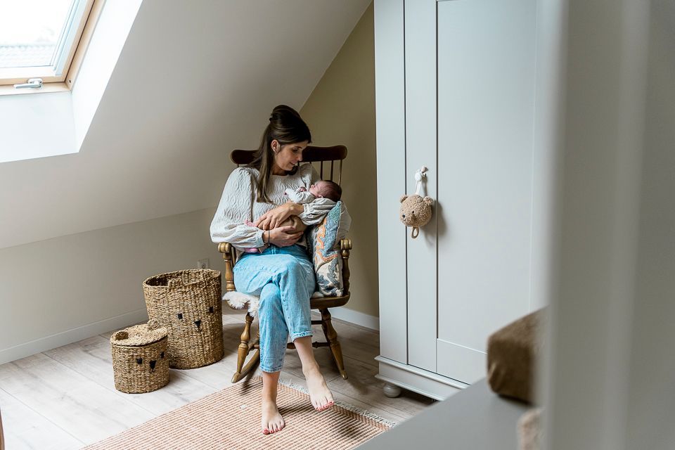 Een vrouw geeft borstvoeding aan een baby in een schommelstoel in een kinderkamer, naast een kledingkast en manden.