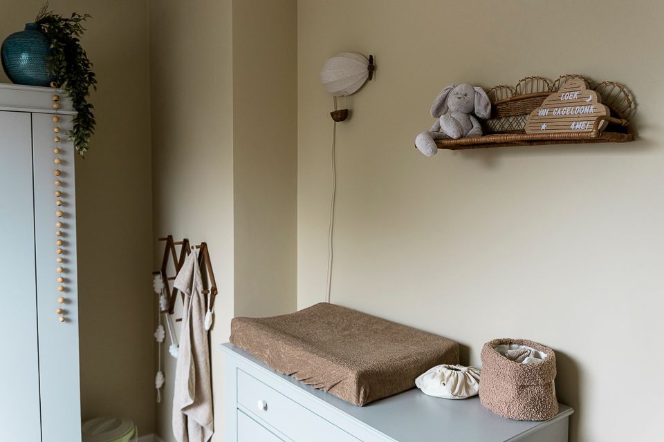 Babyhoekje met commode, aankleedkussen, plankje met konijntje, wandlamp en een blauwe vaas op een wit kastje.