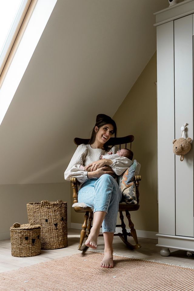 Vrouw in een schommelstoel met een baby in haar armen in een kamer met een kast, rieten manden en een raam.