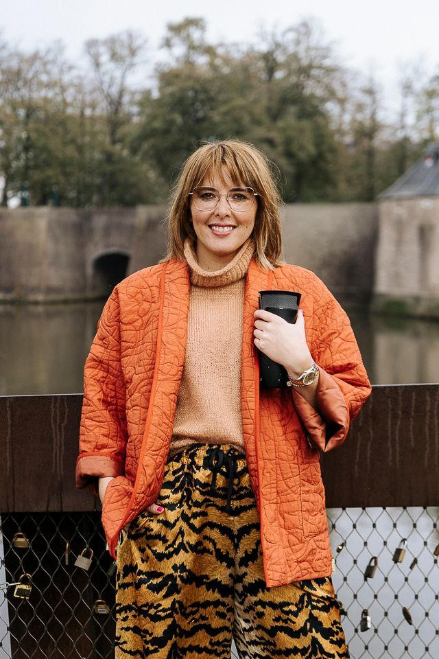 Een vrouw in een oranje jas en een broek met tijgerprint lacht buiten, terwijl ze een zwart voorwerp vasthoudt, met een waterloop en een stenen muur op de achtergrond.