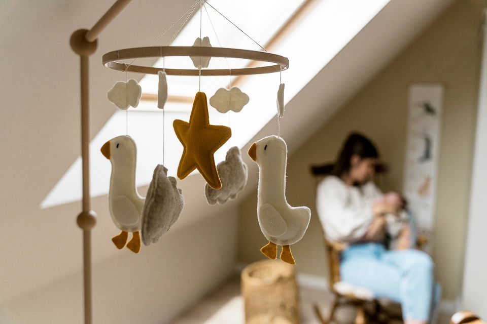 Een babymobiel met ganzen en een ster hangt in een kinderkamer; op de achtergrond is een persoon te zien.