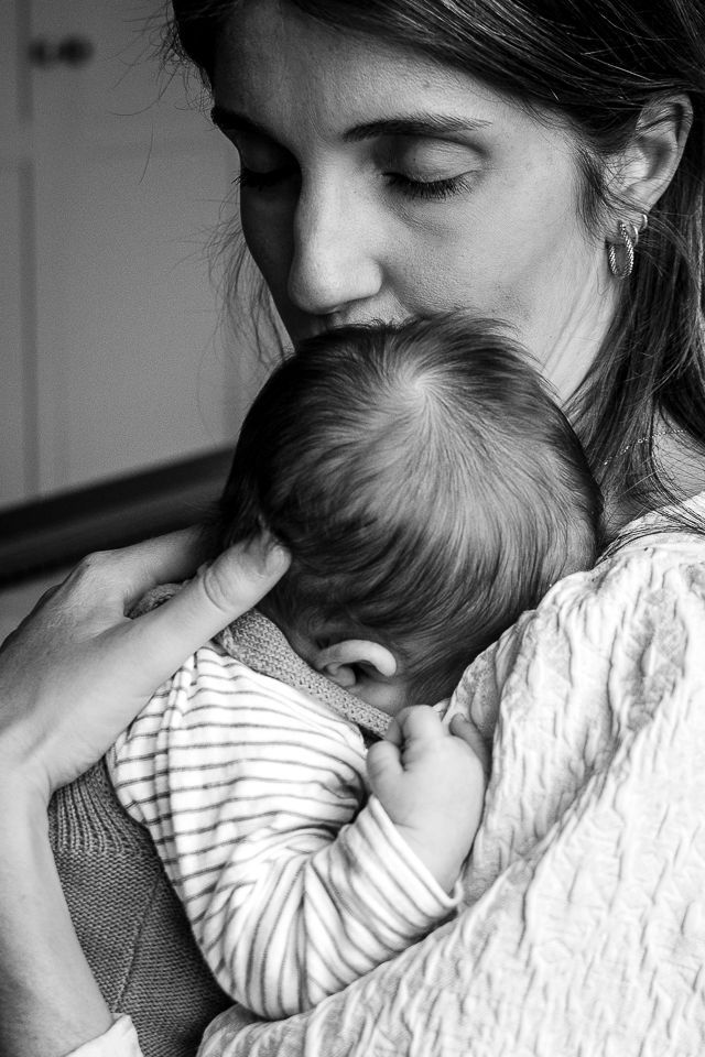 Vrouw houdt een baby vast en kust hem. Het hoofdje van de baby rust op de schouder van de vrouw, die een gestreept shirt draagt. Zwart-wit.
