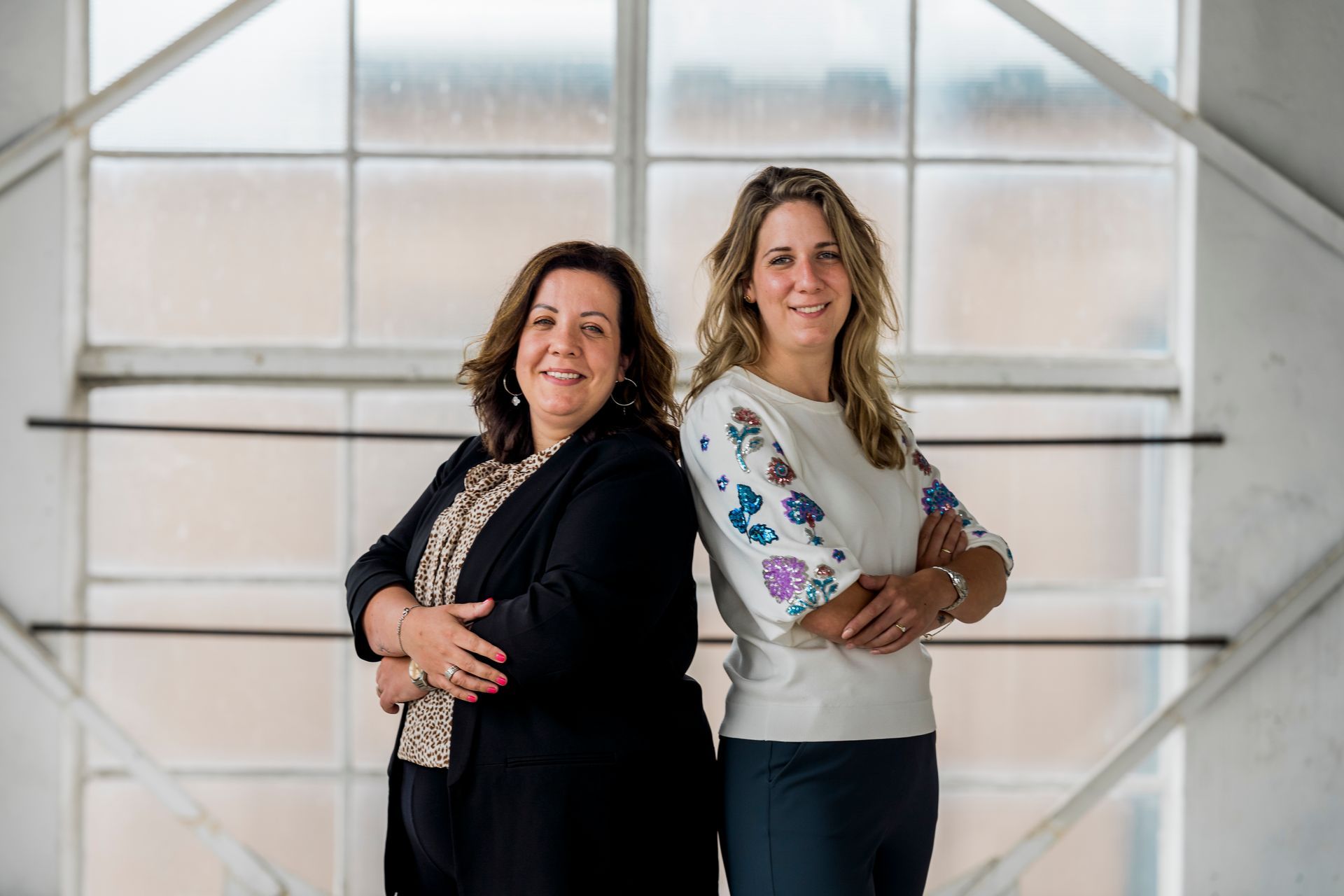 Twee vrouwen staan ​​met hun armen over elkaar voor een raam. De ene draagt ​​een blazer, de andere een blouse met een patroon.
