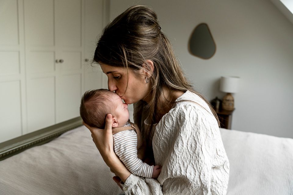 Een vrouw kust het voorhoofd van een baby. Ze liggen binnen op een bed.