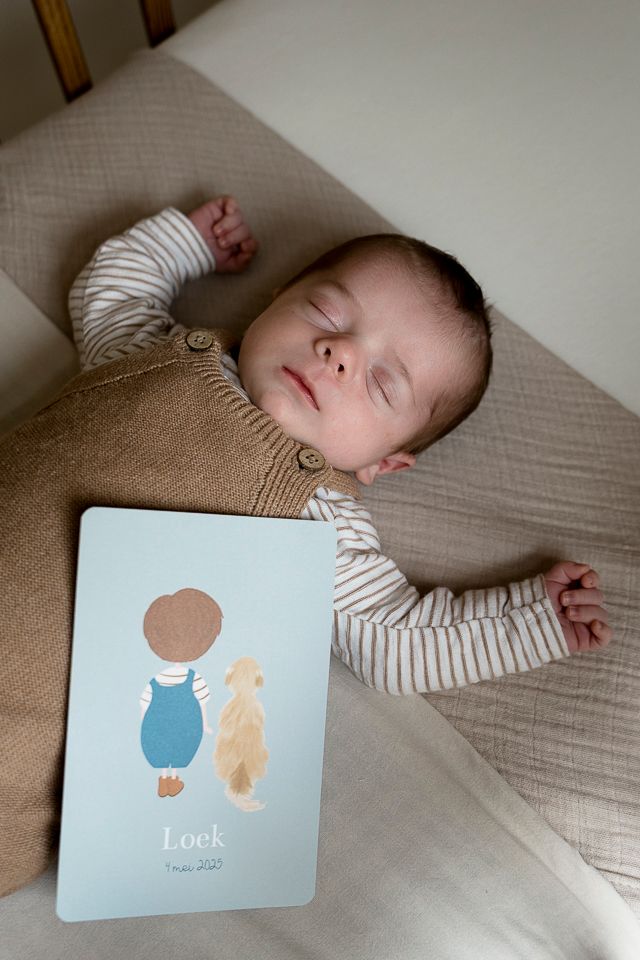 Pasgeboren baby slaapt in de wieg, draagt een bruine overall en een gestreept shirt, met een kaartje ernaast.