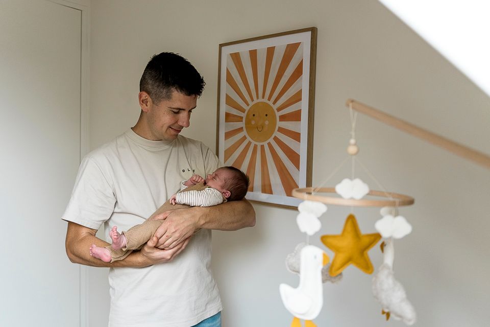 Man met een baby in zijn armen, kijkt naar beneden. Zonkunst en een mobiel met een baby aan de muur. Lichte kamer.