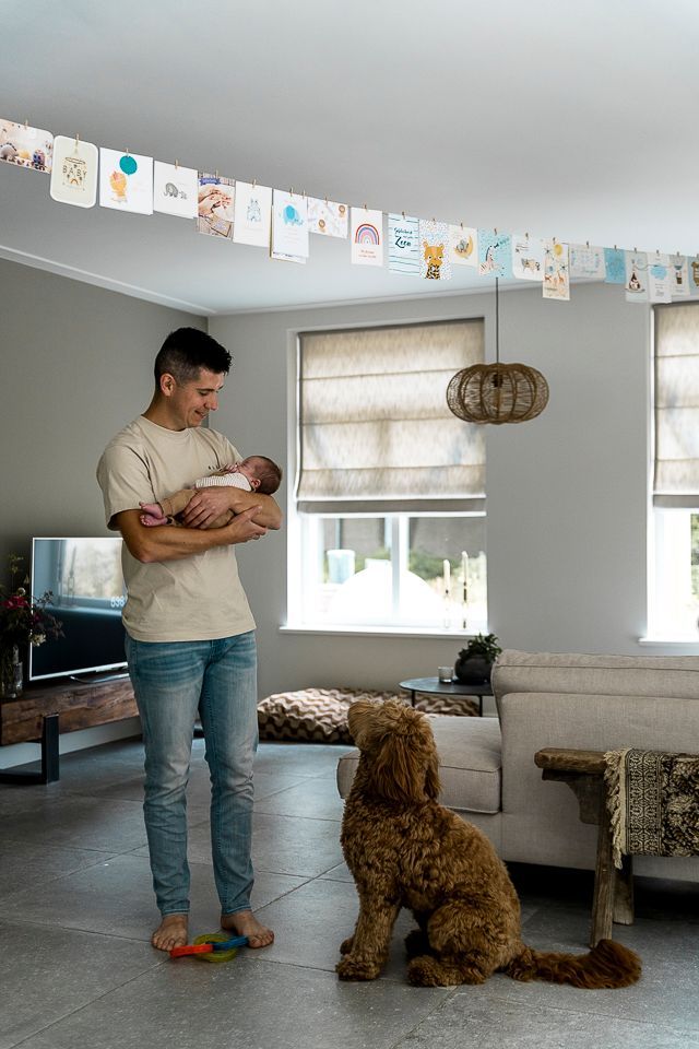 Een man houdt een pasgeboren baby vast, een hond kijkt naar hen in een woonkamer met kaarten aan het plafond.