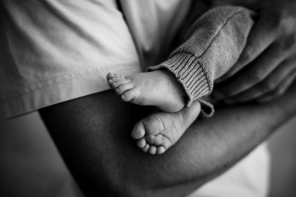 Babyvoetjes en -been in een grijze gebreide broek, vastgehouden in de armen; zwart-wit.