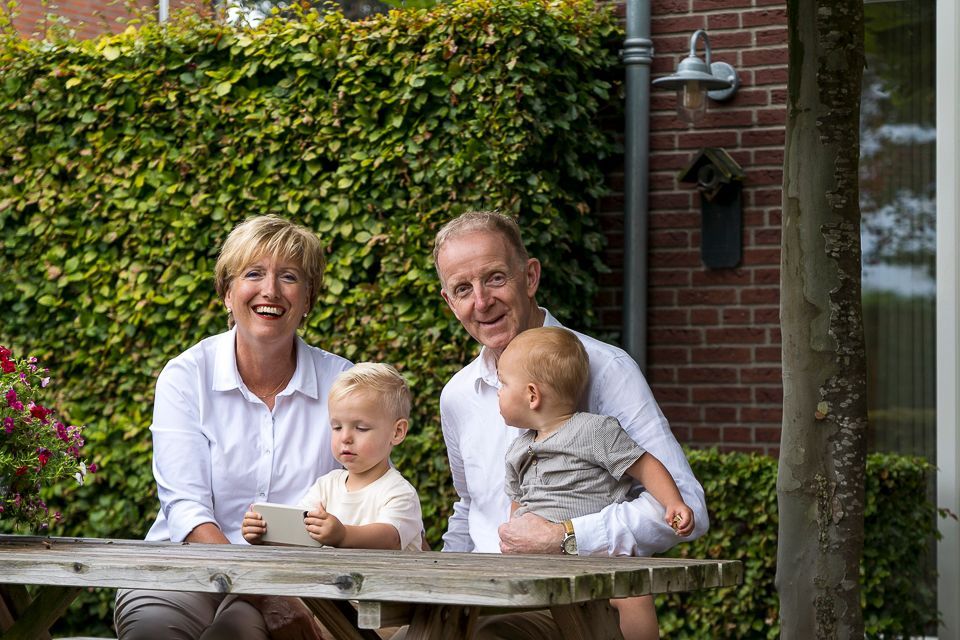 Een gezin met twee jonge kinderen zit aan een houten tafel in de tuin. Op de achtergrond is groen gebladerte te zien en ze glimlachen.