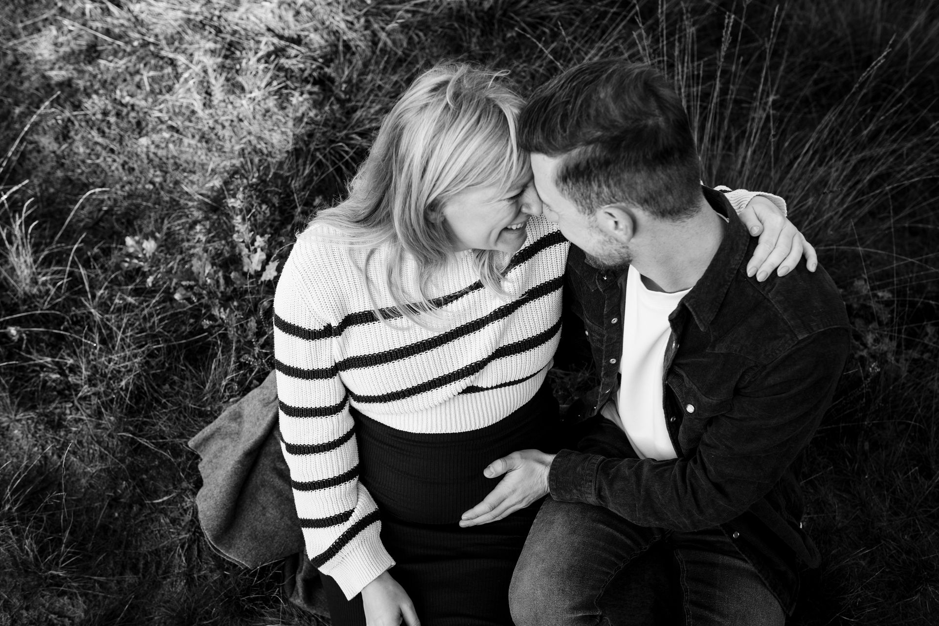 Een stel omhelst elkaar, de zwangere vrouw raakt haar buik aan. Buiten, zittend in hoog gras, in zwart-wit.