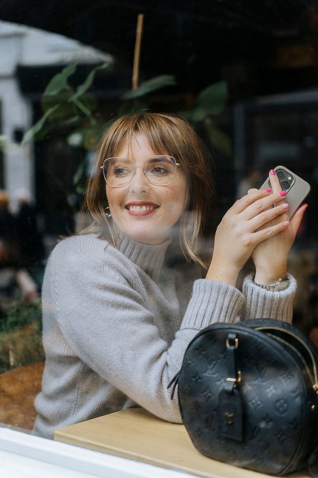 Vrouw met bril, glimlachend en met een telefoon in haar hand, zit bij het raam met een handtas en planten.