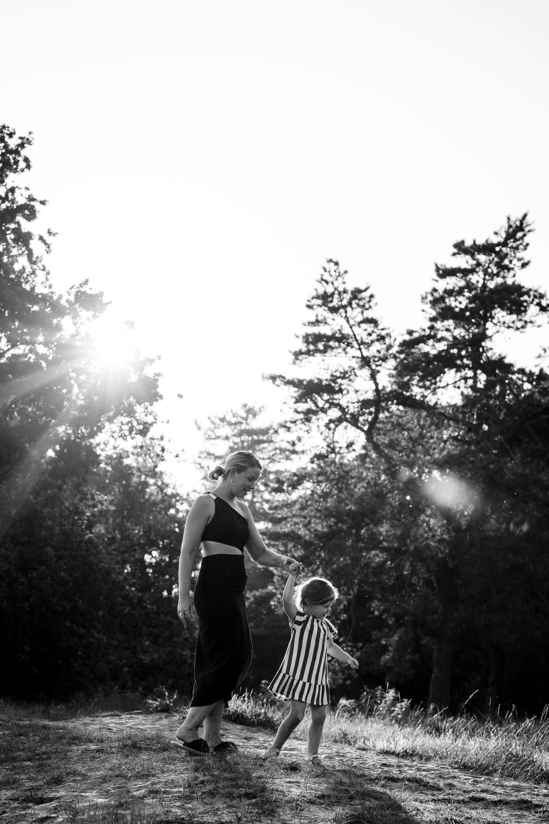 Vrouw en peuter in het bos, hand in hand. De zon schijnt door de bomen en werpt schaduwen.