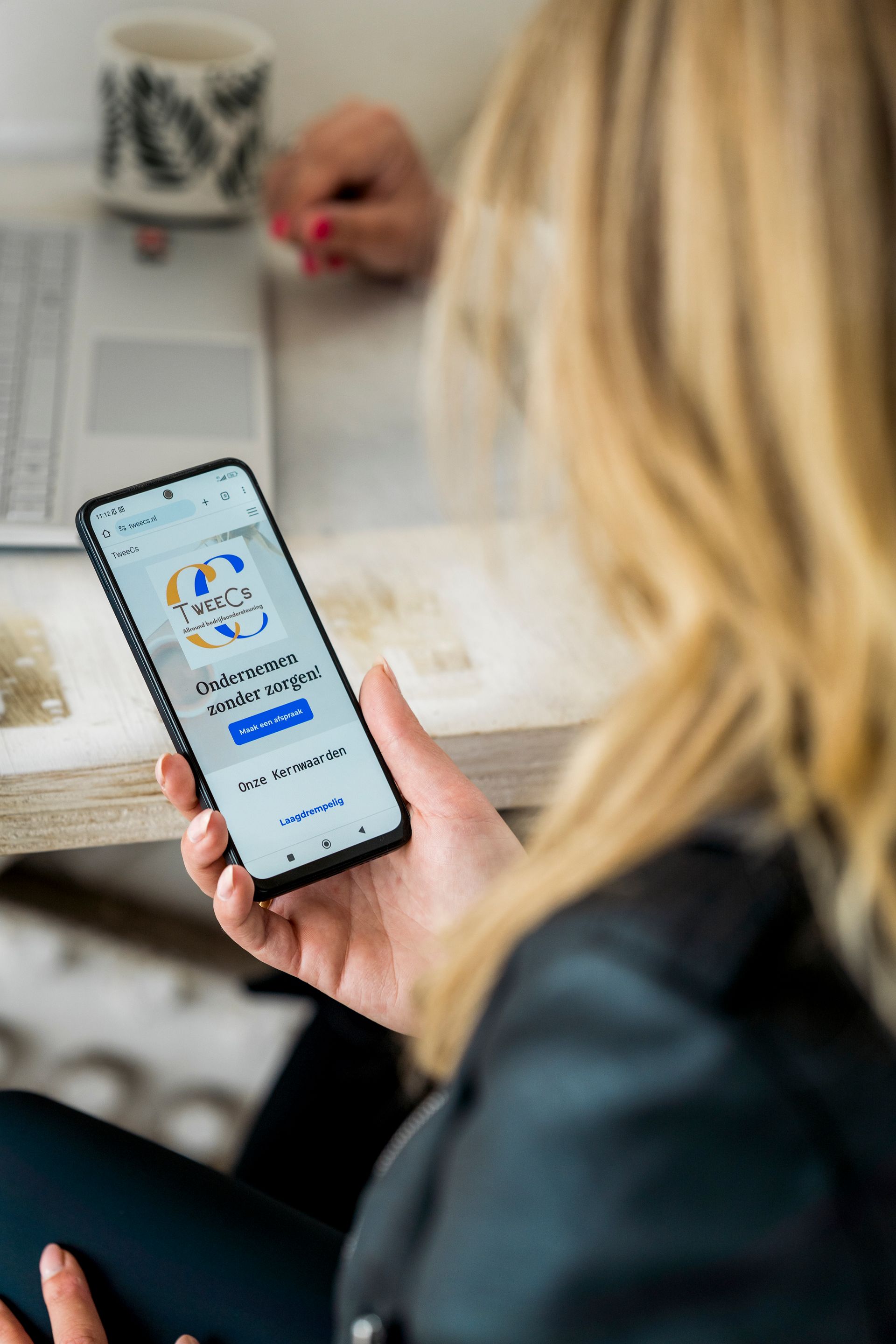 Vrouw met een smartphone waarop een app te zien is; wazige achtergrond met een laptop en een mok.