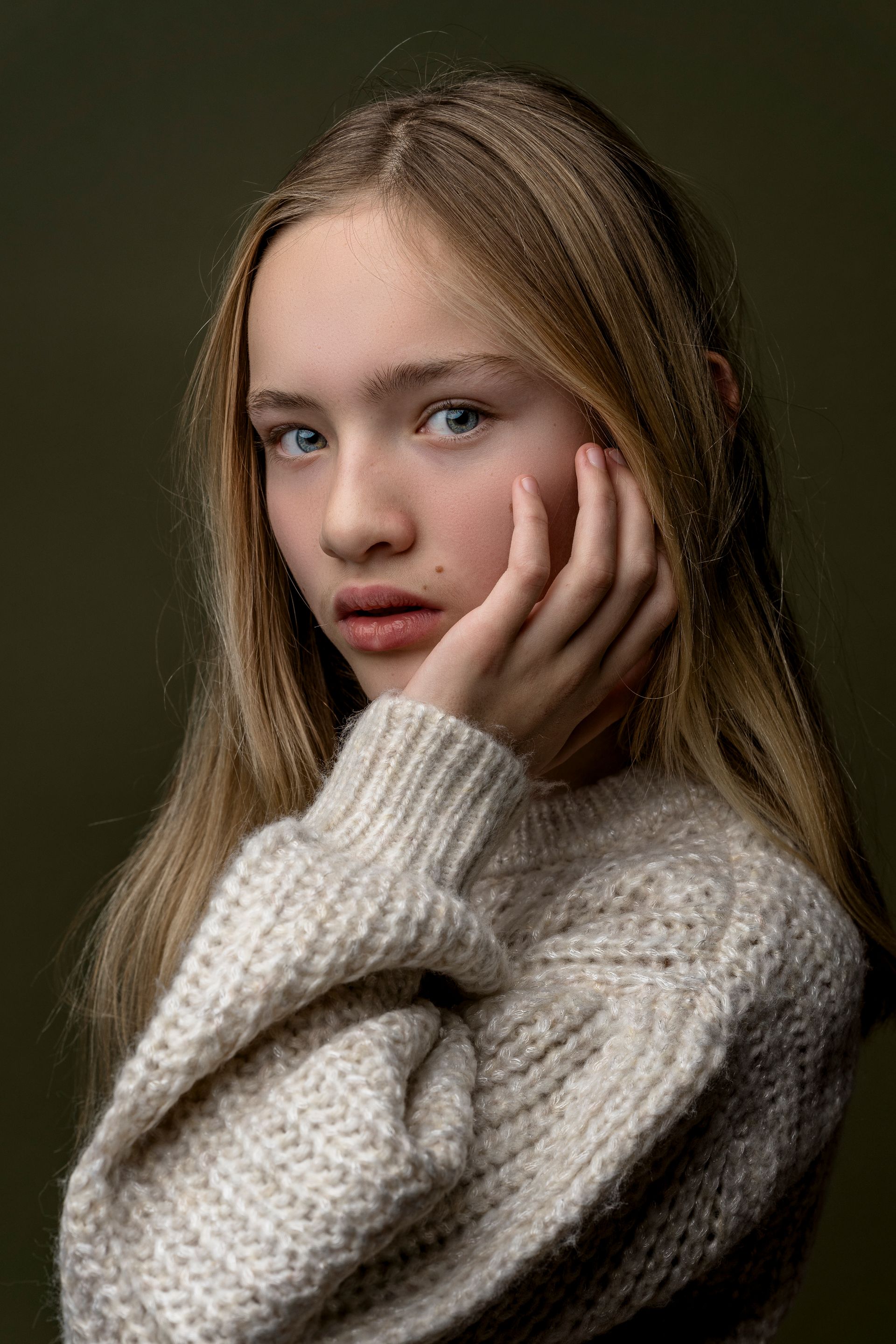 Een jong persoon met lang blond haar, een lichtgekleurde trui, legt een hand op zijn wang en kijkt de kijker recht aan, tegen een groene achtergrond.