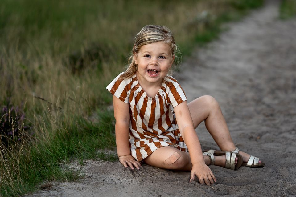 Een meisje in een gestreepte jurk lacht terwijl ze op een zandpad zit.