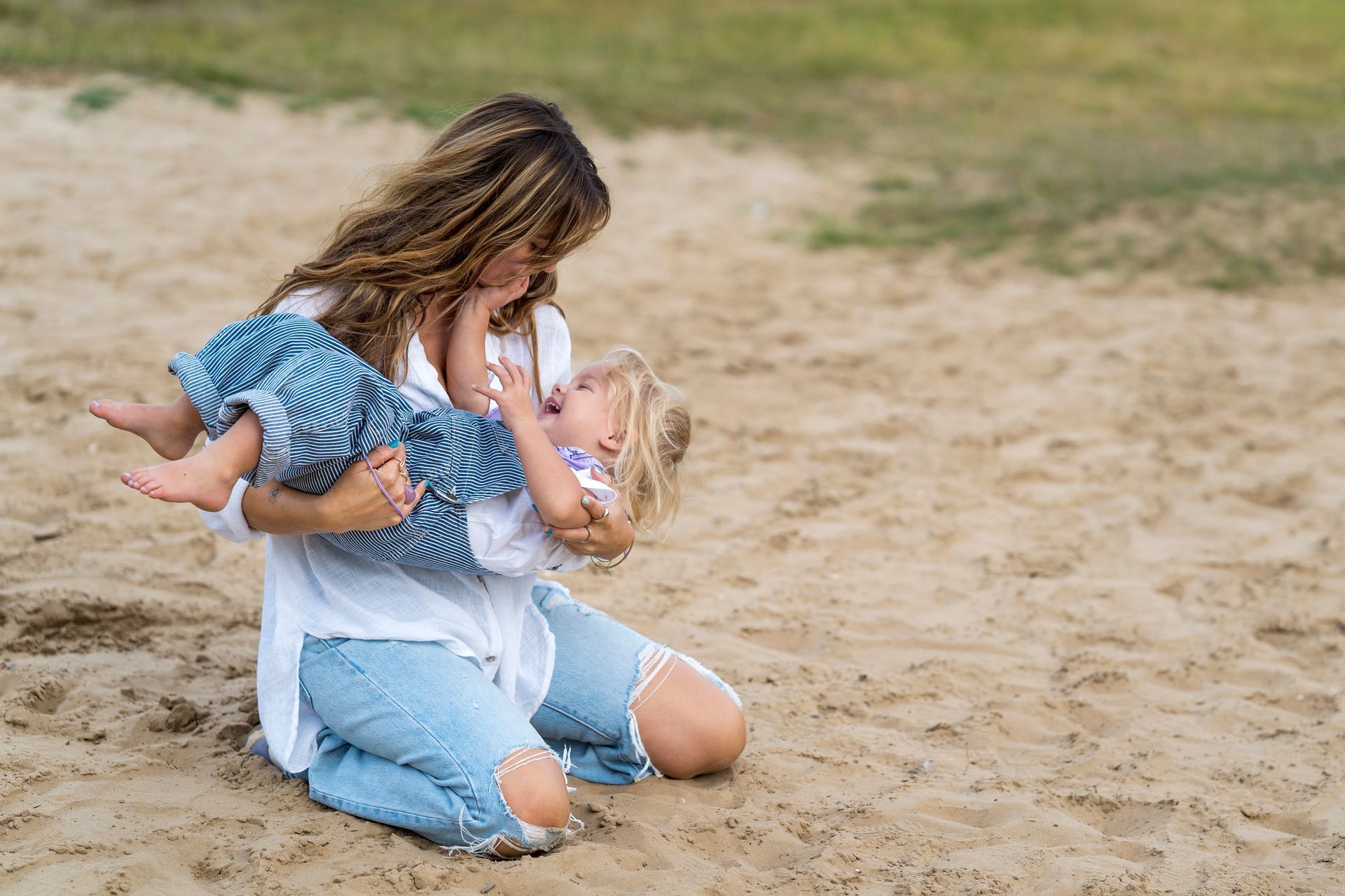 Vrouw knielt in het zand, houdt een lachend kind omhoog en kijkt ernaar; buiten.