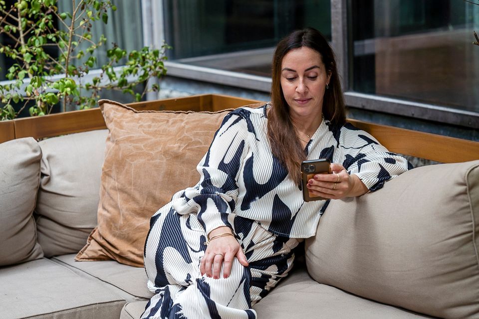 Vrouw zit op een bank en kijkt naar haar telefoon. Ze draagt ​​een zwart-wit gestreepte jurk, er liggen bruine en beige kussens omheen, en de foto is buiten genomen.
