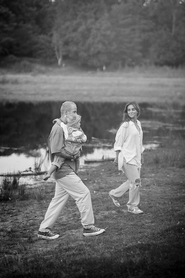 Een gezin wandelt langs een waterpartij. Een man draagt ​​een kind in een draagzak; een vrouw loopt ernaast. Op de achtergrond is een bos te zien.