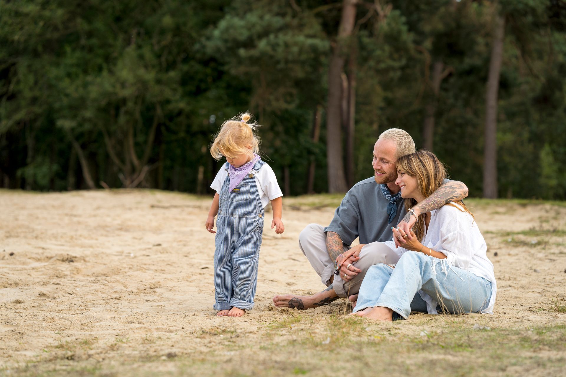Een gezin zit in het zand. Een kind in een overall staat er vlakbij. Op de achtergrond zijn bomen te zien.