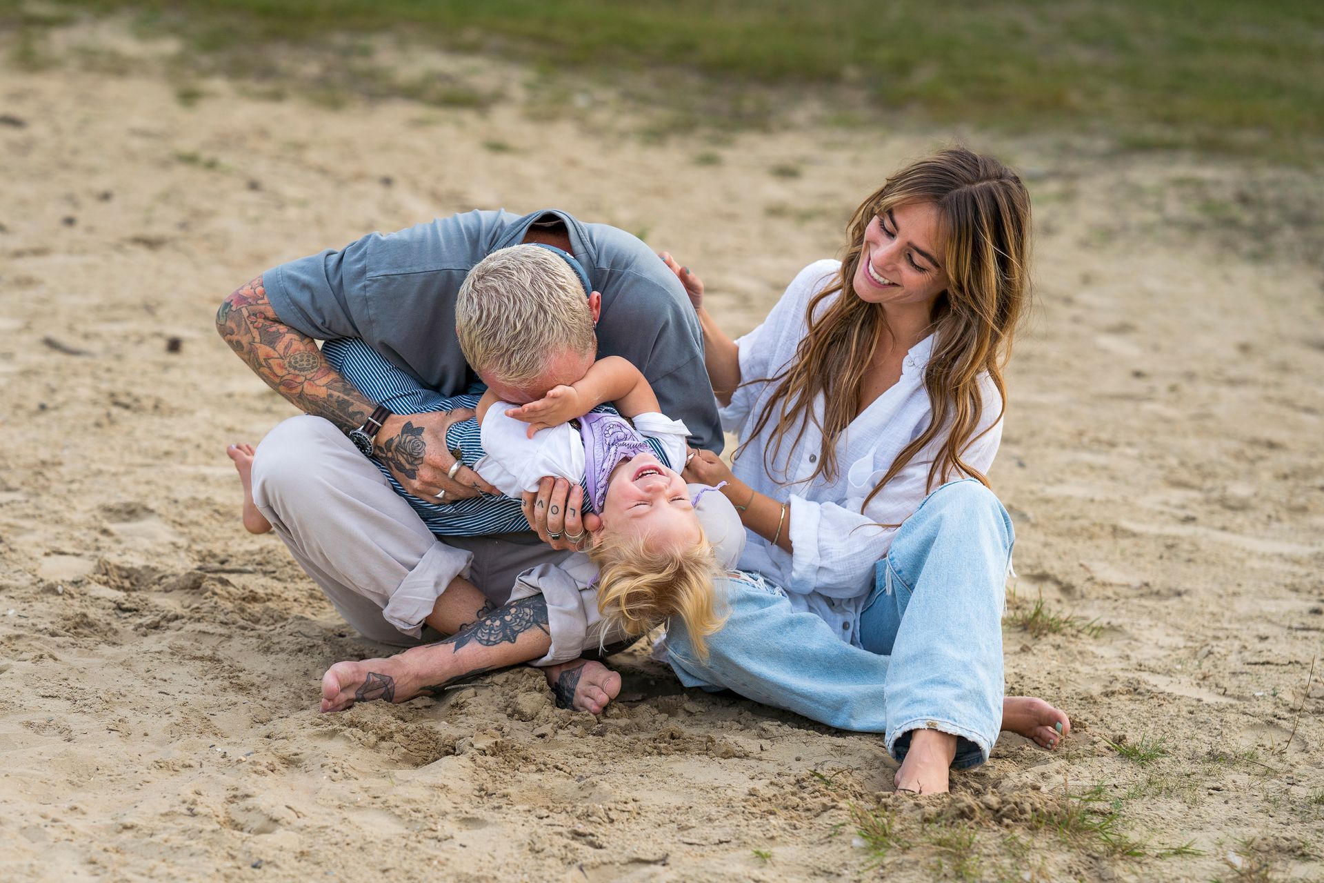 Familie speelt in het zand: vader kietelt kind, moeder kijkt glimlachend toe. Blond haar, tatoeages, lichte kleding.