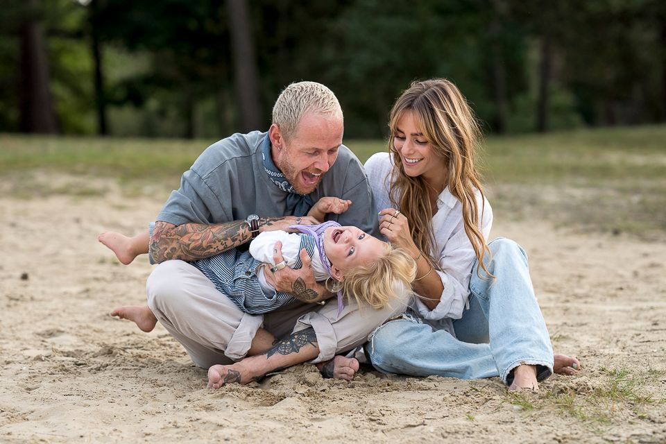 Een gezin lacht en speelt in het zand; een man met tatoeages, een vrouw en een kind.