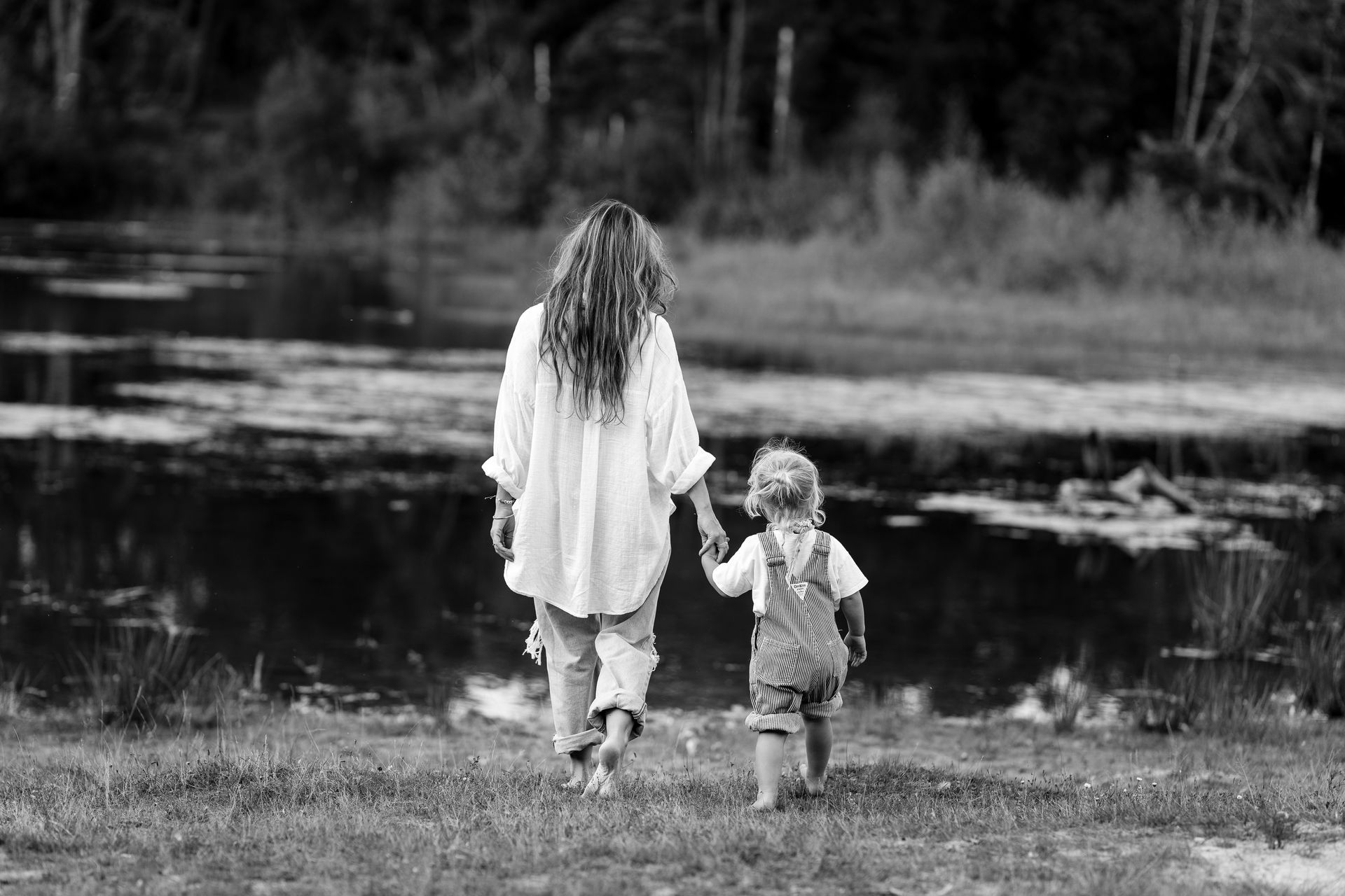 Een vrouw in een witte blouse en een kind in een overall lopen hand in hand naar een waterpartij.