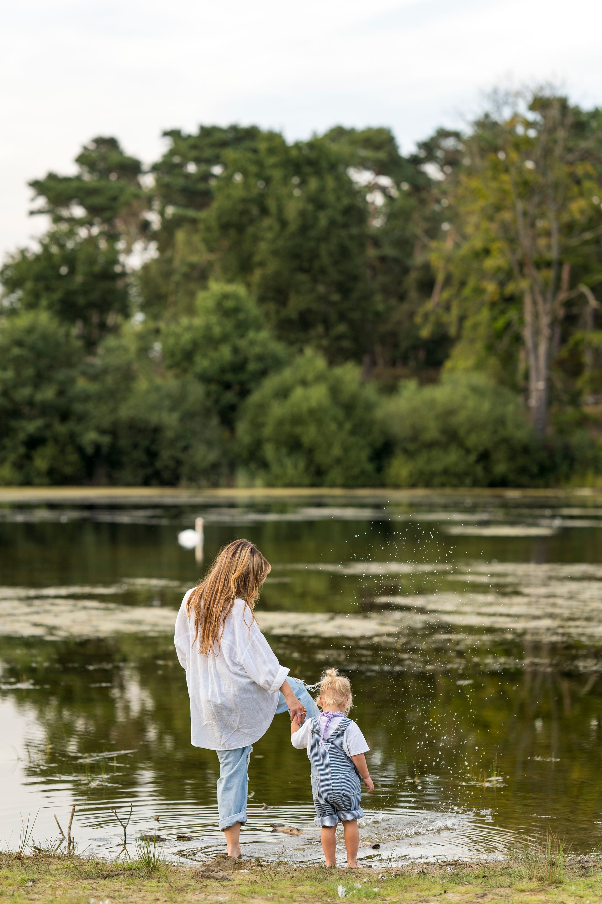 Vrouw en kind spelen bij een meer. Ze staan in het ondiepe water en spetteren. Op de achtergrond is een bos te zien.