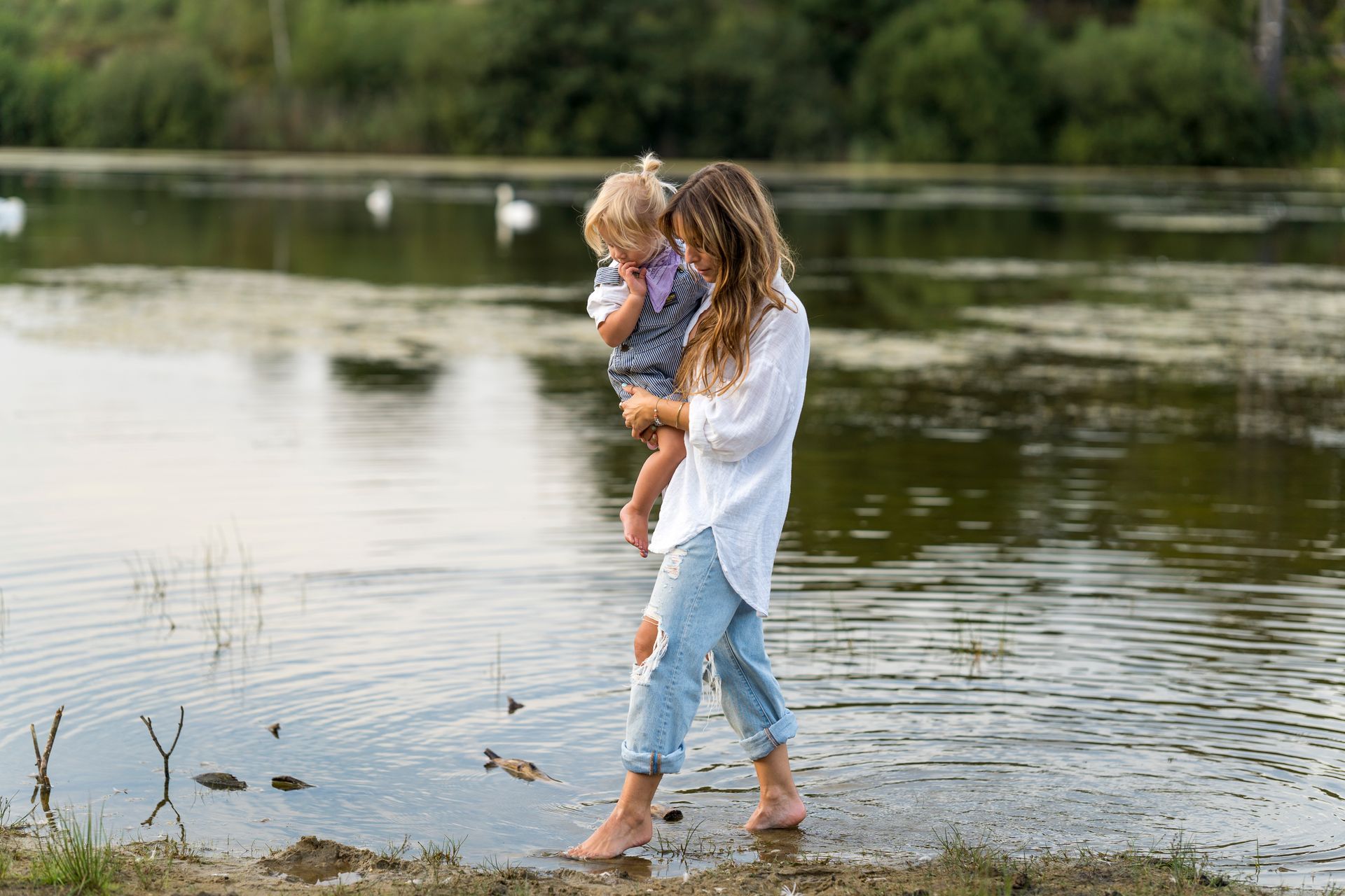 Een vrouw met een kind in haar armen waadt in een meer. Op de achtergrond zwemmen zwanen in het meer.