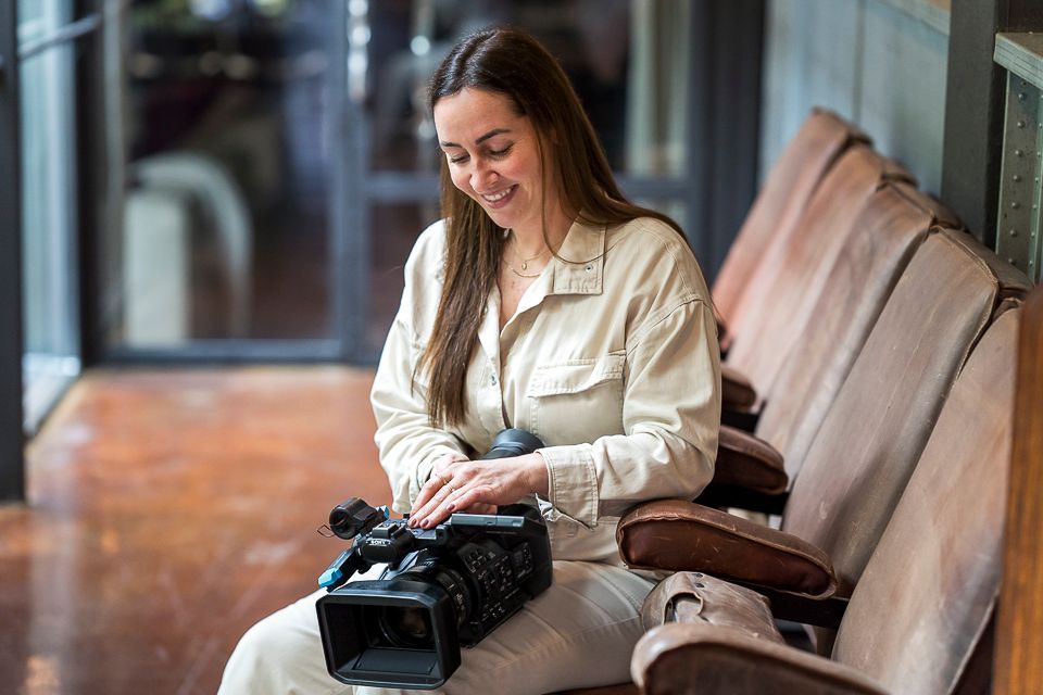 Vrouw in beige outfit glimlacht terwijl ze een videocamera afstelt, zittend op een houten theaterbank.