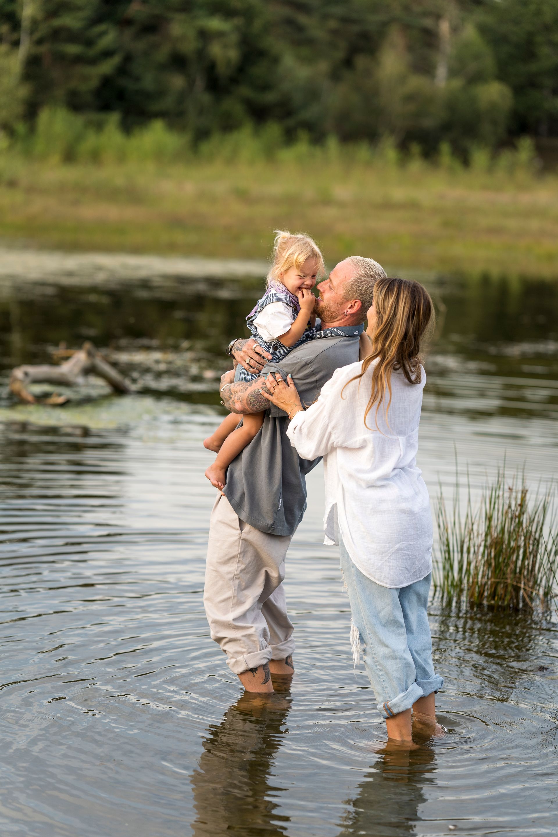 Een gezin waadt door het water; een man houdt een kind vast en kust het, een vrouw staat er vlakbij. Op de achtergrond is groen gebladerte te zien.