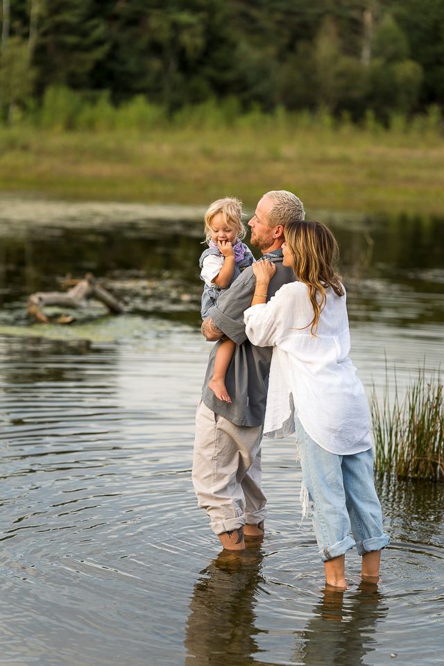 Een gezin waadt door het water. Een man houdt een kind vast; een vrouw omhelst een man. Op de achtergrond is groen gebladerte te zien.