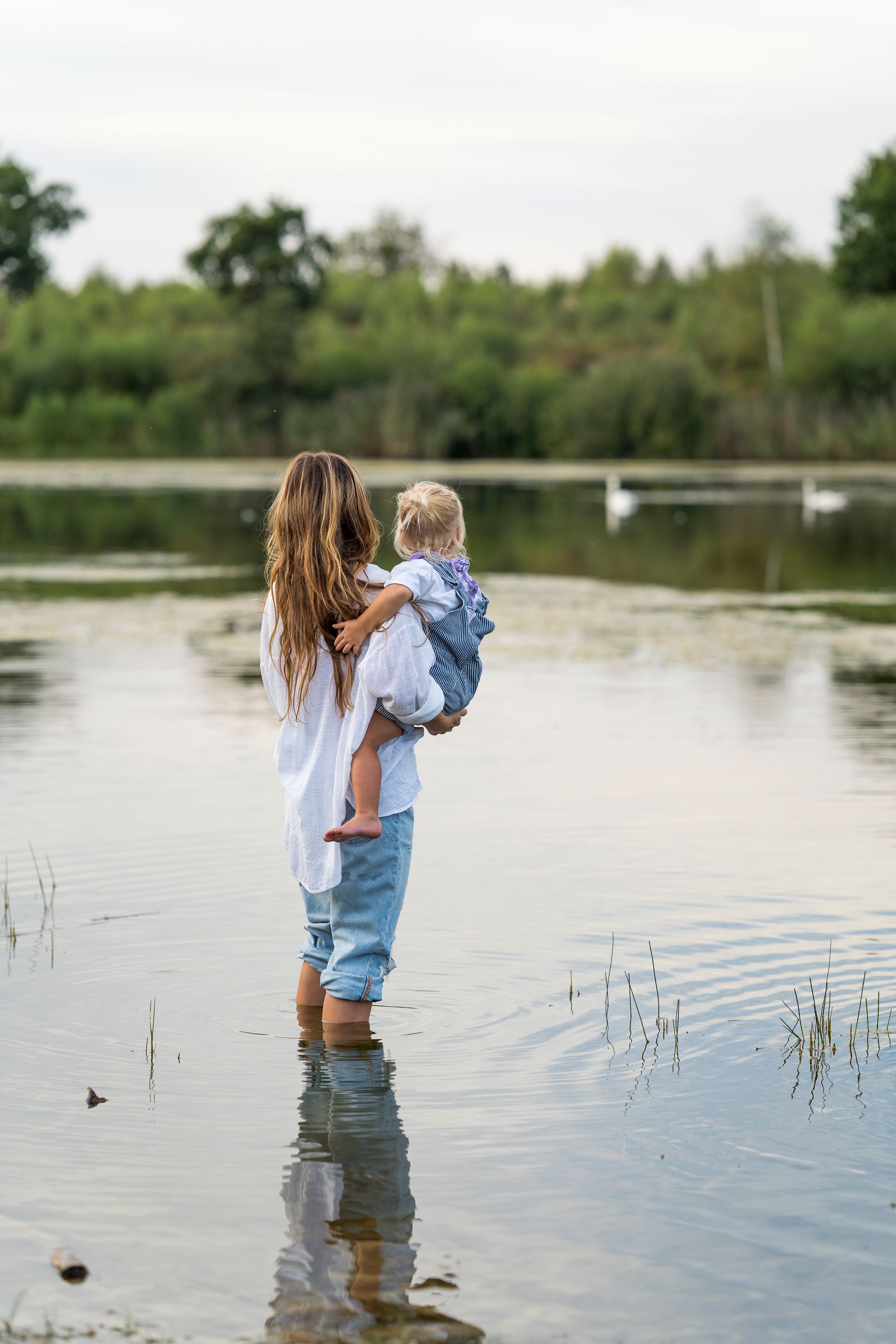 Vrouw houdt kind vast in ondiep water, het beeld van het kind wordt weerspiegeld. Meer en bomen op de achtergrond.