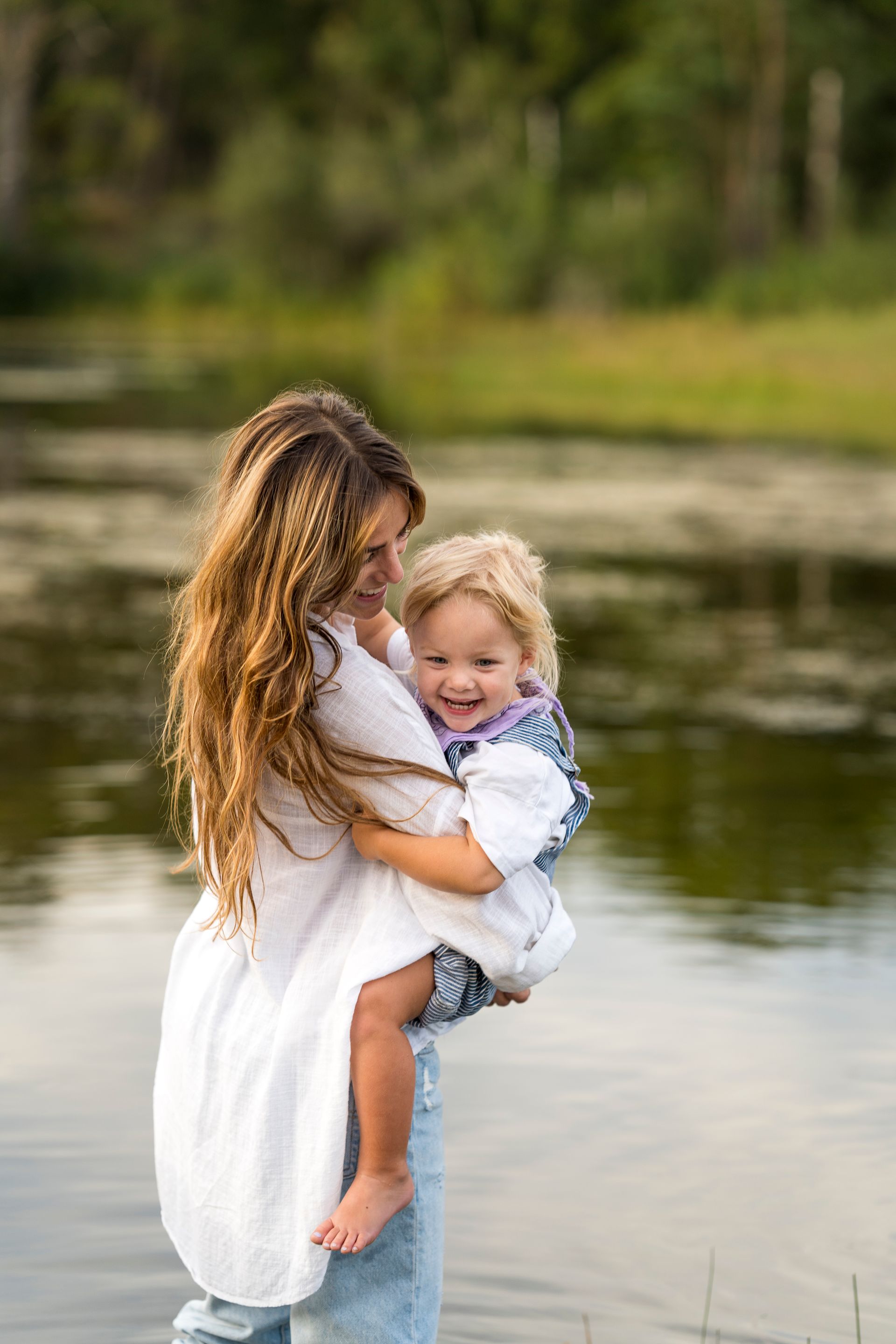 Vrouw in witte blouse houdt lachend kind vast bij het water.