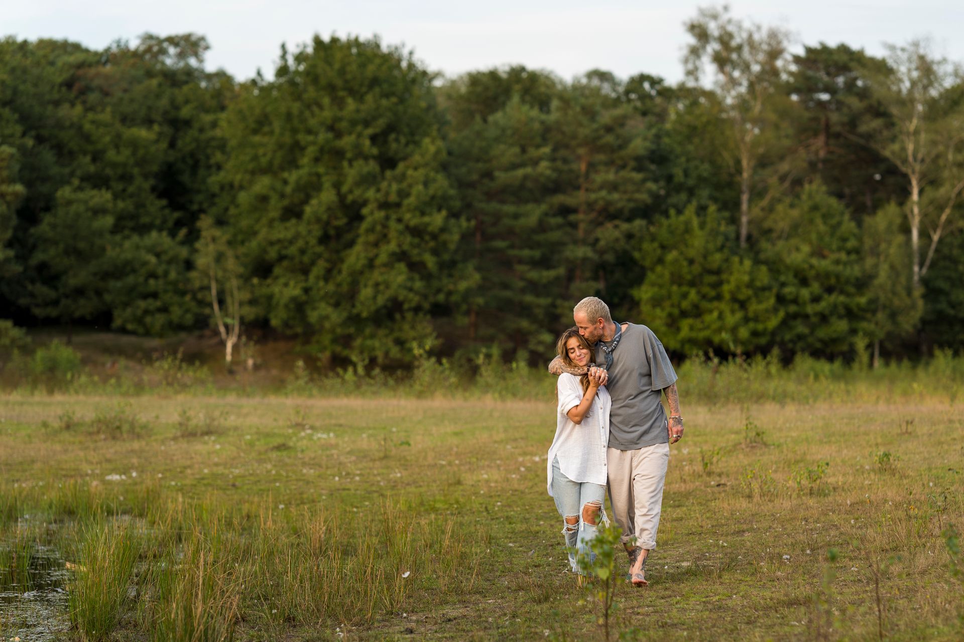 Een stel loopt door een veld, de man heeft zijn arm om de vrouw heen geslagen, met een bos op de achtergrond.