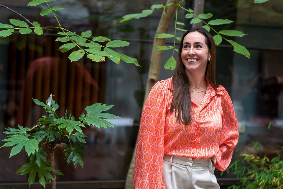 Een vrouw in een oranje blouse en crèmekleurige broek lacht buiten, tussen groene planten.