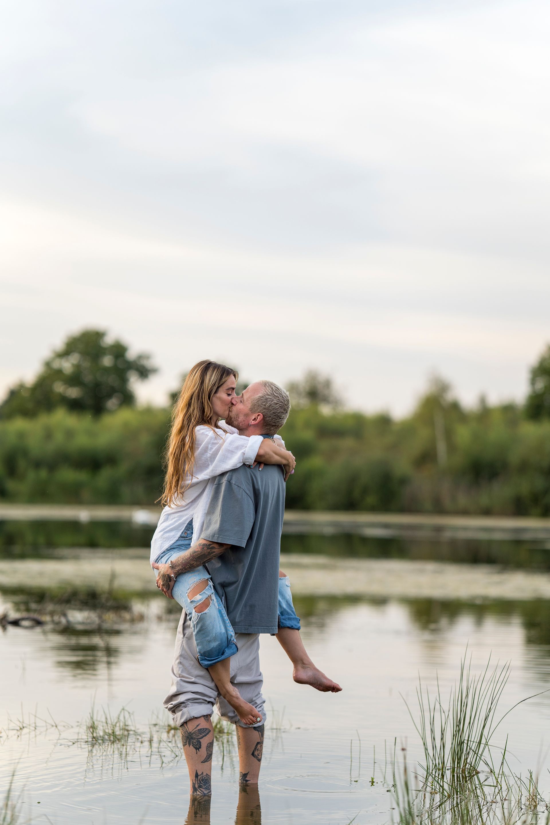 Een man draagt een vrouw en ze kussen elkaar in ondiep water. Lichtblauwe lucht, groene bomen op de achtergrond.